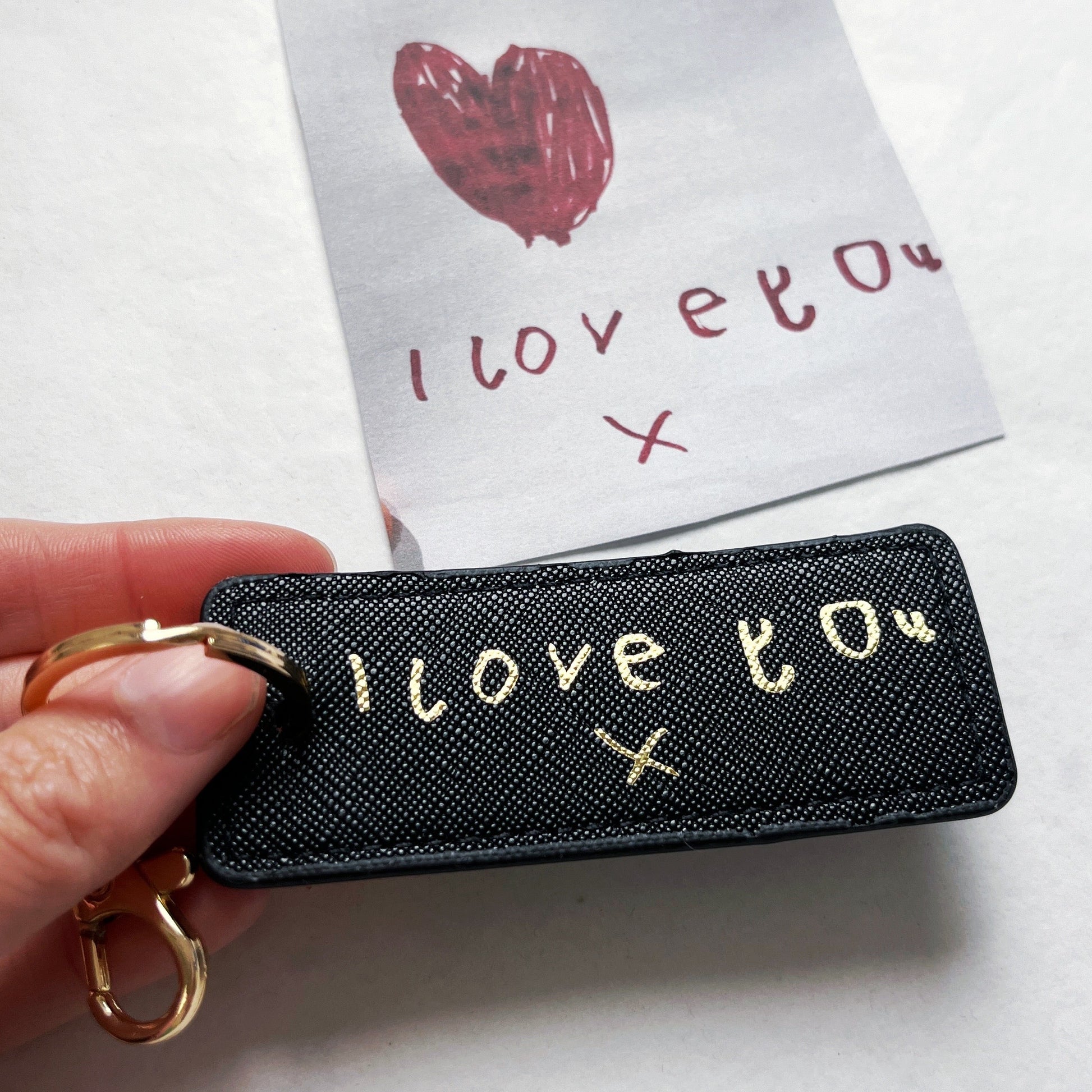 Black keychain with 'I love you' text held by a hand, with a small card in the background.