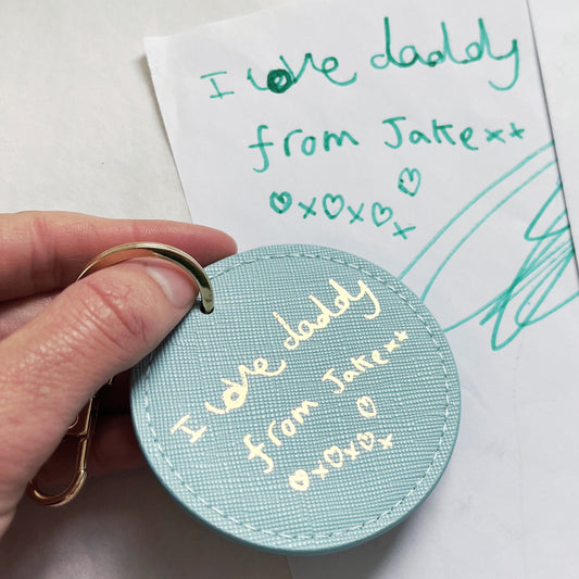 Round blue keychain with 'I love daddy' text held over a handwritten note on a white background