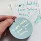 Round blue keychain with 'I love daddy' text held over a handwritten note on a white background